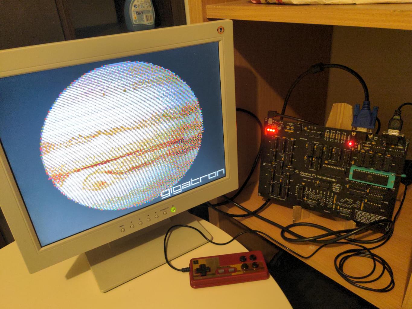 A picture of a white LCD monitor on the left and a populated PCB on the right connected to it; between those there is a small Famicom like controller; there is a picture of Jupiter on the screen and the word "Gigatron".